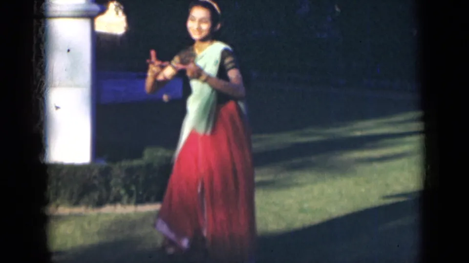 In 1947 India footage, a woman in a saree appears to smile while forming a dance gesture near shrubs and a white pillar.