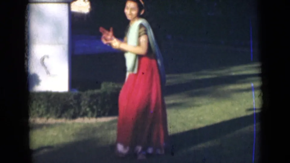 In 1947 India footage, the dancer turns partly sideways with hands extended while standing near a white pillar and garden shrubs.
