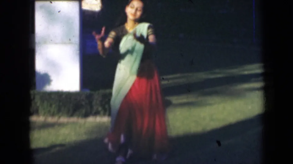 In 1947 India home-movie footage, a woman in a red and pale green sari dances outdoors near a white pillar and hedge.