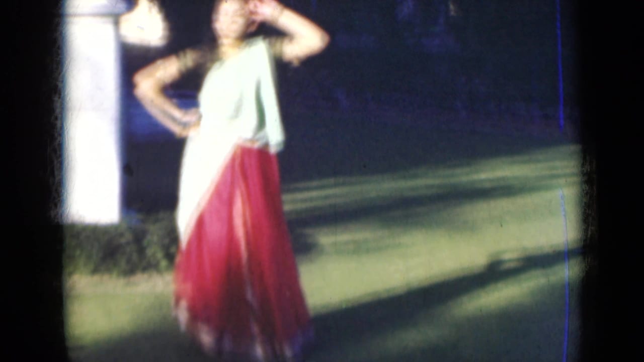 The dancer poses with one hand near her head, her red skirt and pale drape bright against the shaded lawn.