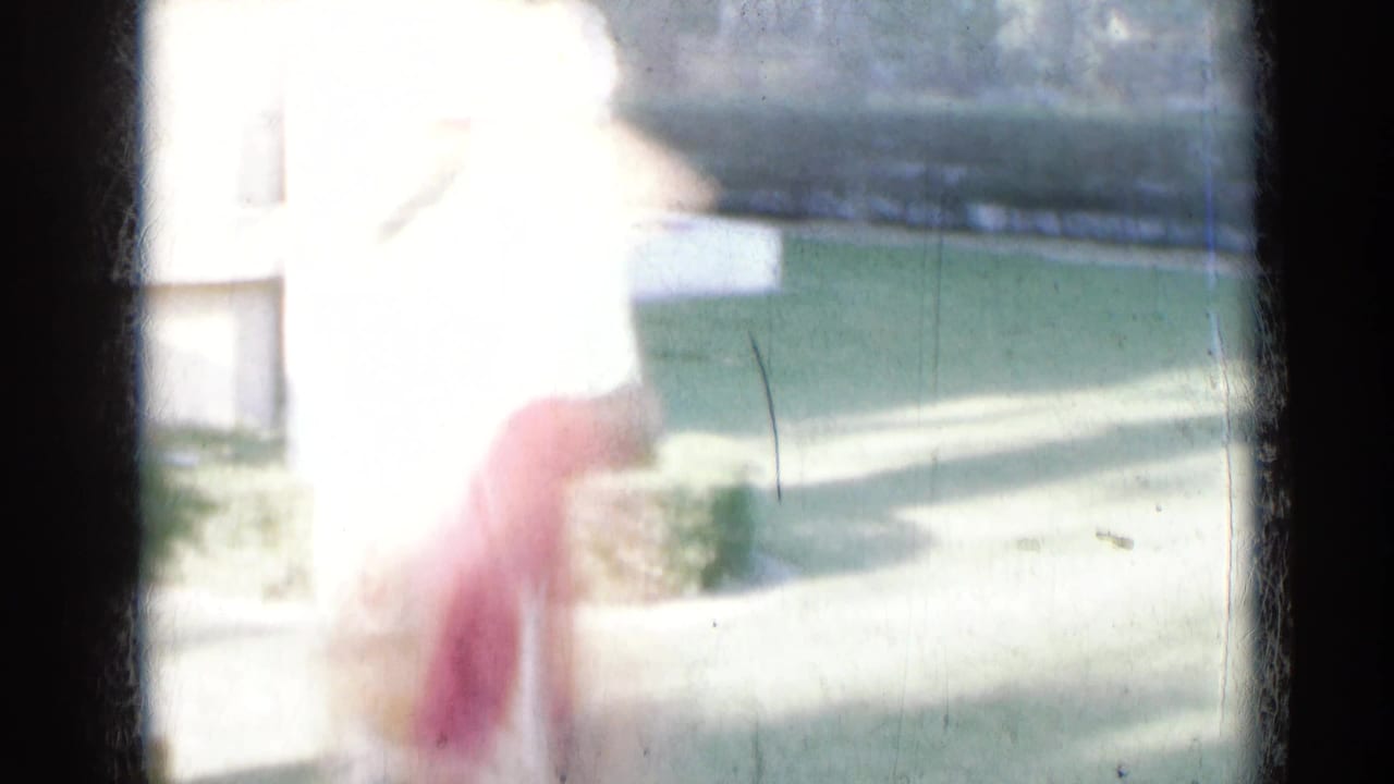 Overexposed 8mm frame from India showing a woman in red and green clothing moving outdoors near a lawn and pale building.