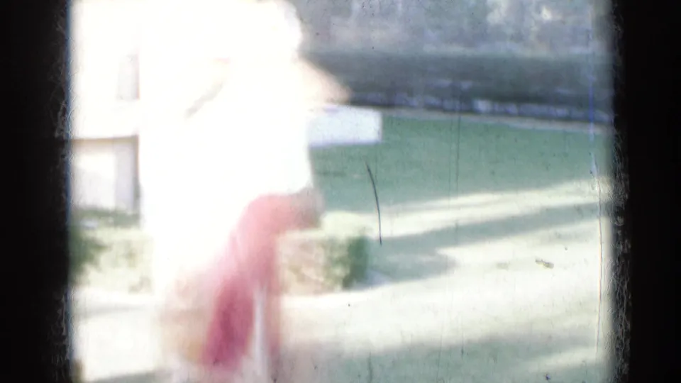 Overexposed 8mm frame from India showing a woman in red and green clothing moving outdoors near a lawn and pale building.