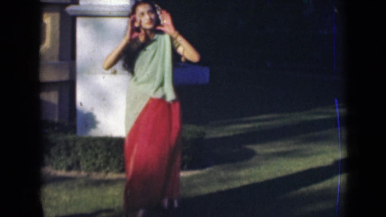 In 1947 India home-movie footage, a woman in a green and red sari raises both hands while dancing beside a white column and lawn.