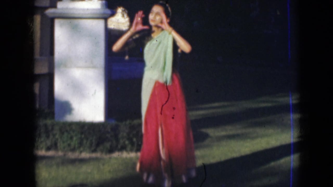 A woman in a green and red sari smiles toward the camera with raised dance gestures beside a white pillar in India, 1947.