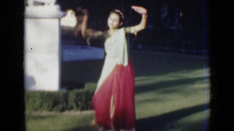 A 1947 India 8mm frame shows the woman extending one arm upward in a dance pose near a white column, hedge, and lawn.
