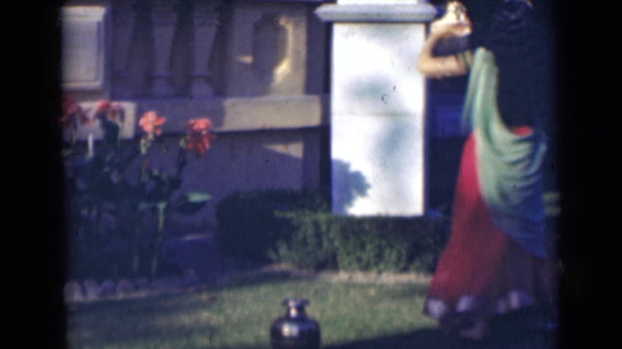 In supported 1947 India footage, a woman in a red and green sari turns in a garden beside a white pillar and flowering plants.