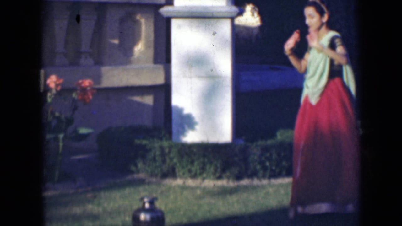 A woman in a red and green sari gestures with both hands while dancing outdoors in India, with a lawn and white pillar behind her.