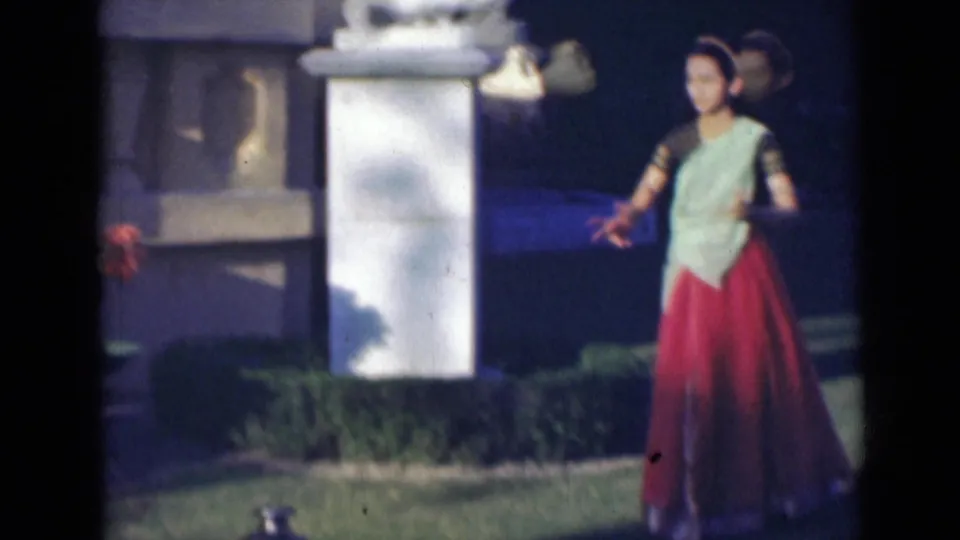 The woman faces the camera with arms extended during the dance, framed by a white pillar, lawn, and flowering plants in the garden.