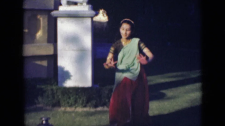 A woman in a red and green sari holds a poised hand gesture while dancing outdoors, with the garden pillar and lawn visible behind her.