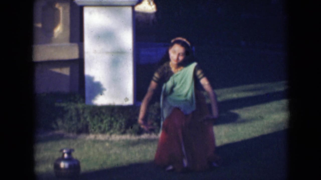In 1947 India footage, a woman in a green and red saree dances on a lawn beside hedges and a pale stone pillar.