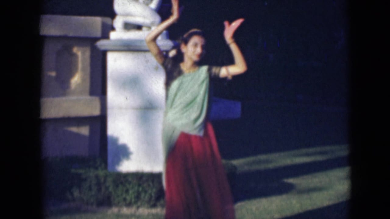 In 1947 India footage, the dancer raises her arms beside a white stone pillar and dark garden background.