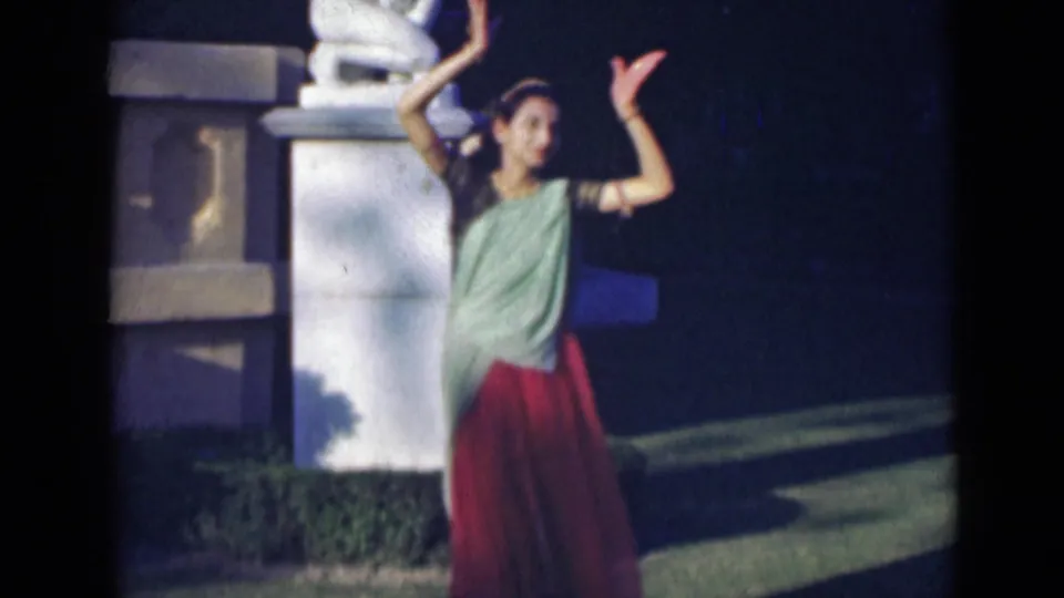 In 1947 India footage, the dancer raises her arms beside a white stone pillar and dark garden background.