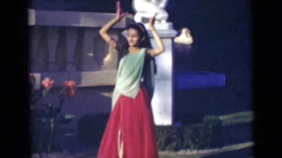 In 1947 India footage, the woman holds a raised-arm dance pose near red flowers, hedges, and a pale stone pillar.