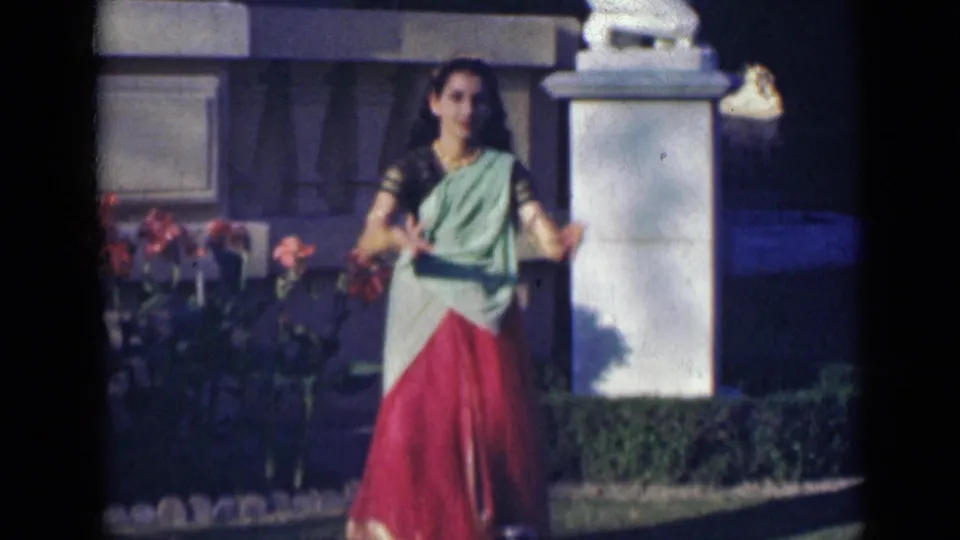 In 1947 India footage, the dancer faces the camera with open hand gestures near flowers, hedges, and stone garden features.