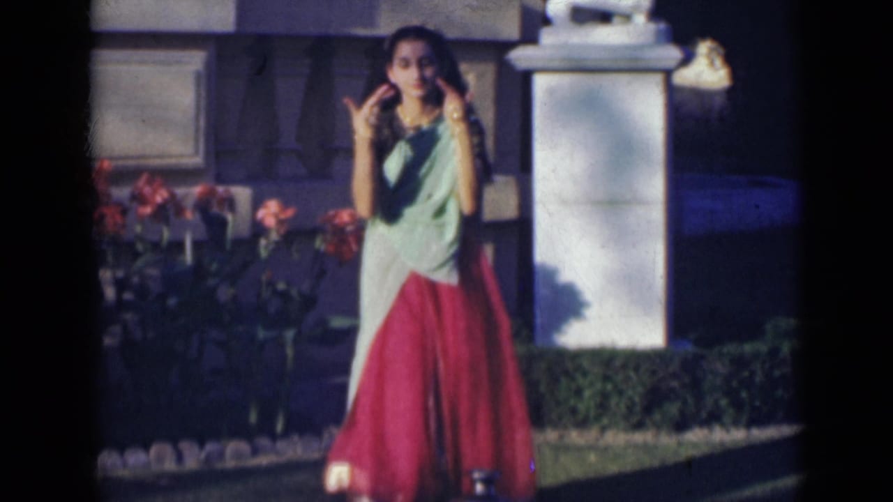 In 1947 India footage, a woman in a red and pale green sari stands near red flowers and a white stone pillar with one hand raised.