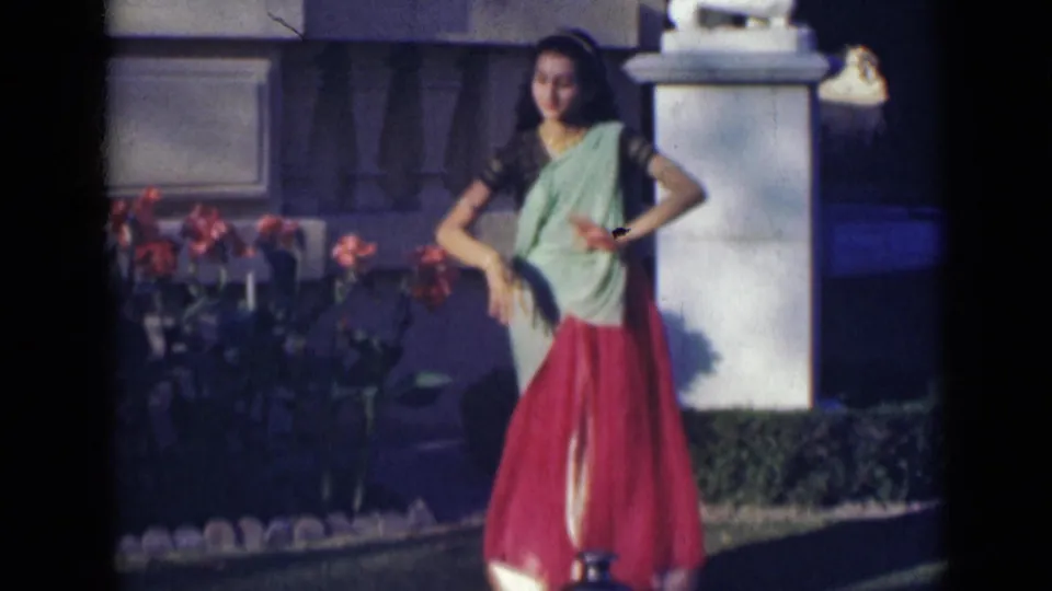 A woman in traditional Indian clothing turns in a garden setting, with flowers, lawn, and a stone pillar visible behind her.