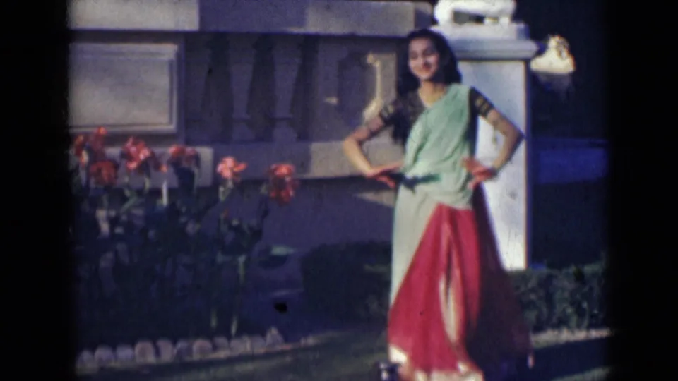 In the final frame, the woman stands smiling in a red and pale green sari near red flowers and a white pillar in India footage from 1947.