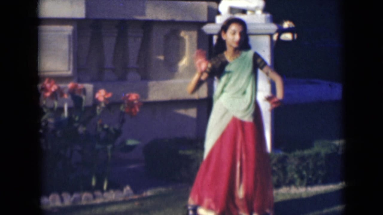 Young woman in green and red clothing dances outdoors beside red flowers and a stone garden structure.