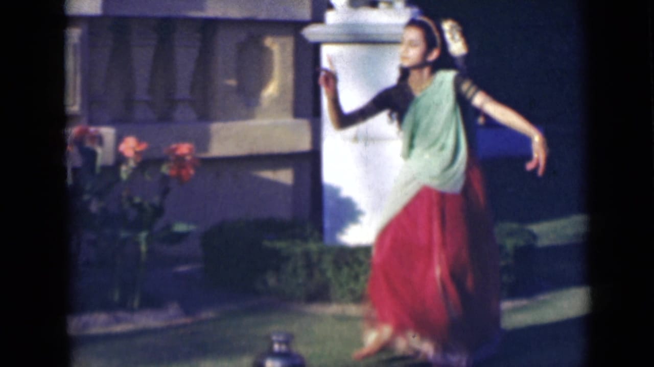 Young woman dances on grass with arms raised near a garden wall and flowering plants.