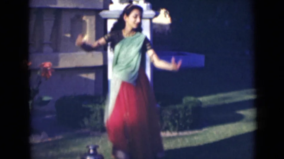 A young woman in a green drape and red skirt continues her dance in a garden, captured with soft 8mm color and grain.
