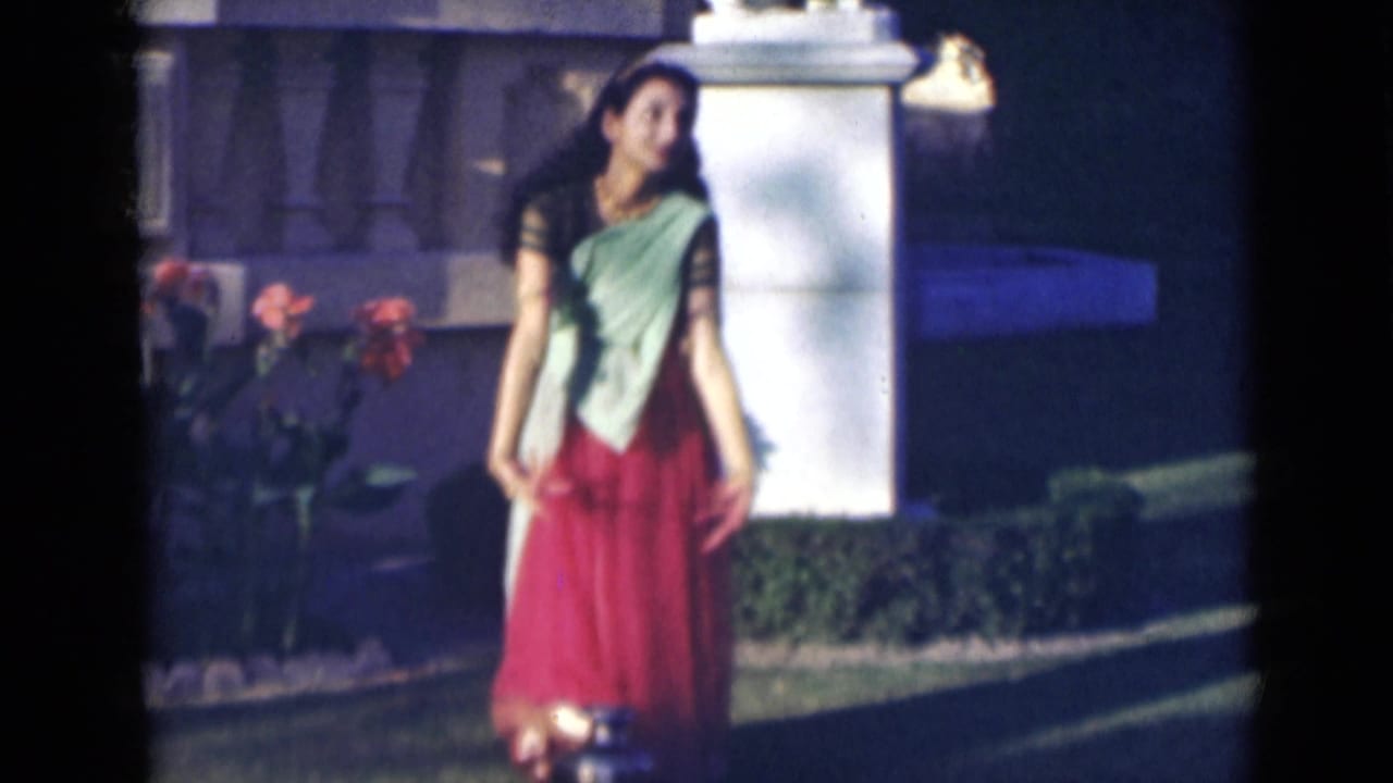 Woman in a red and light green sari standing in a garden near flowers and a white pillar.