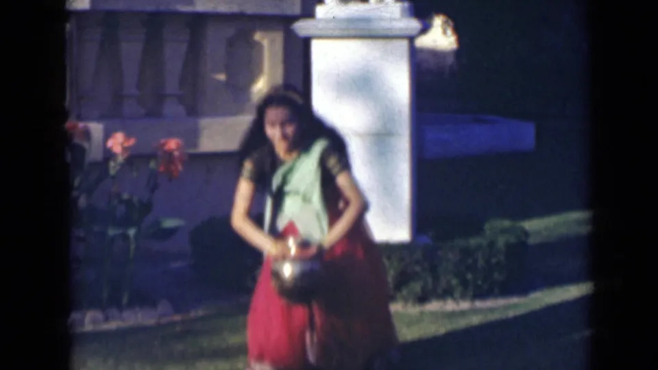 In 1947 India, the woman bends forward in her sari as she handles a small metallic vase on the garden lawn.