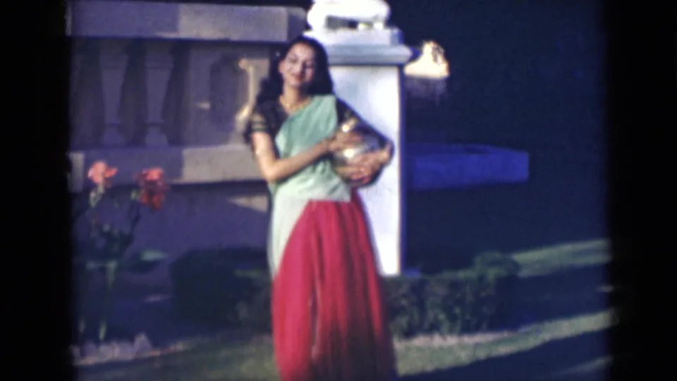 In 1947 India, the woman stands in the garden holding a small metallic vase against her sari near hedges and a white pillar.