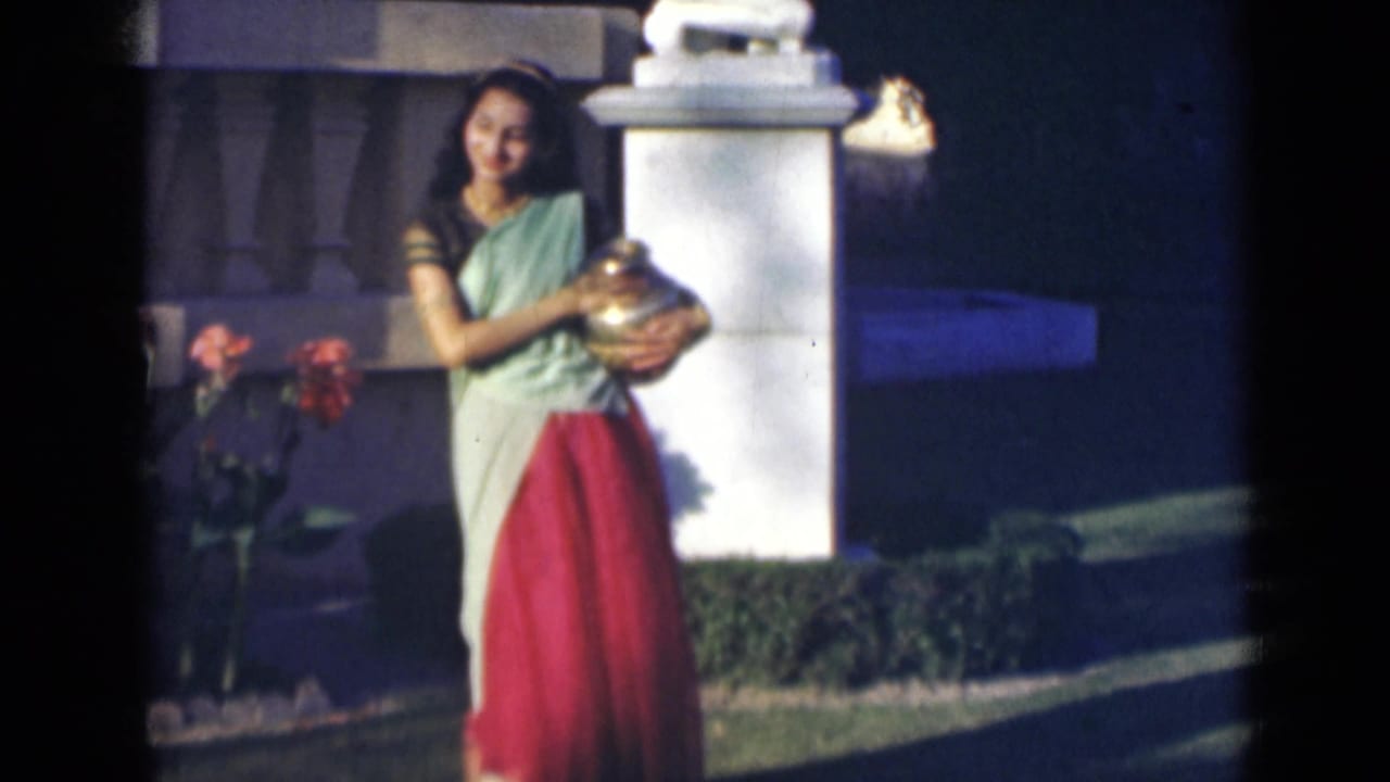 Young woman in a light green and red outfit holds a large metallic vessel in a garden.