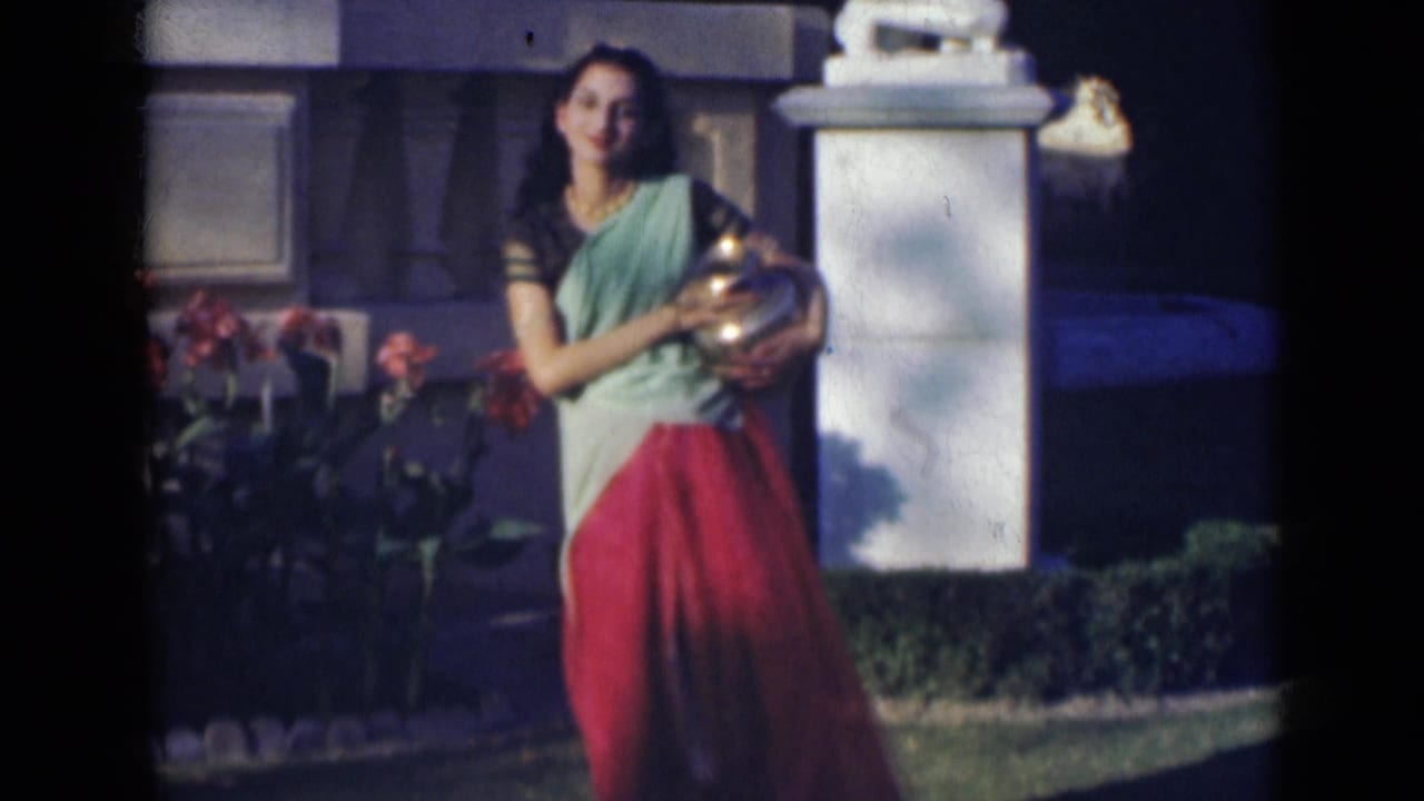The woman shifts her pose with the metal vessel held close, framed by flowers, lawn, and a pale garden structure.