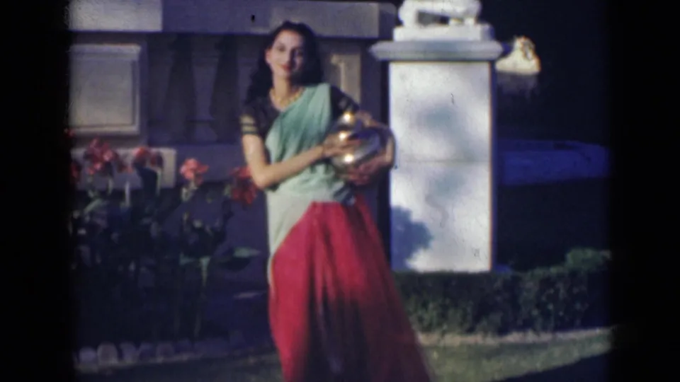 The woman shifts her pose with the metal vessel held close, framed by flowers, lawn, and a pale garden structure.
