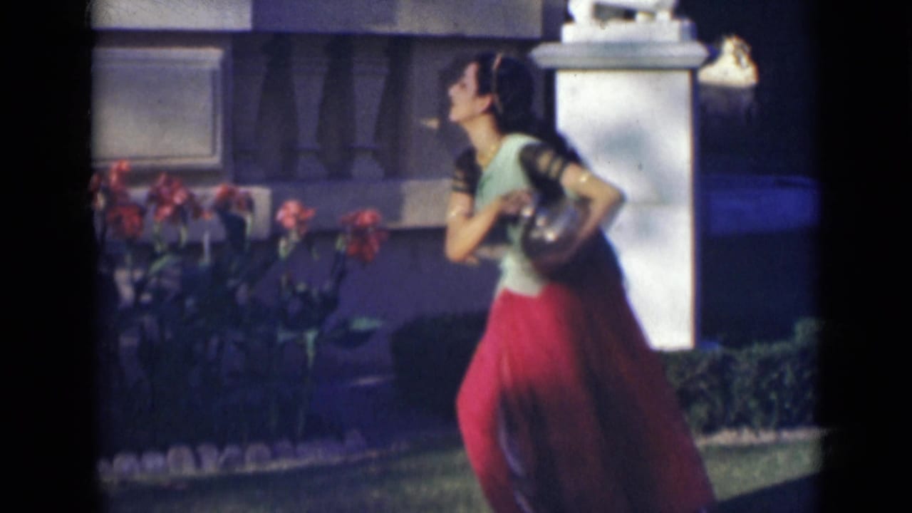 The woman turns partly in profile while holding the vessel, with flowers, grass, and a white garden pillar behind her.