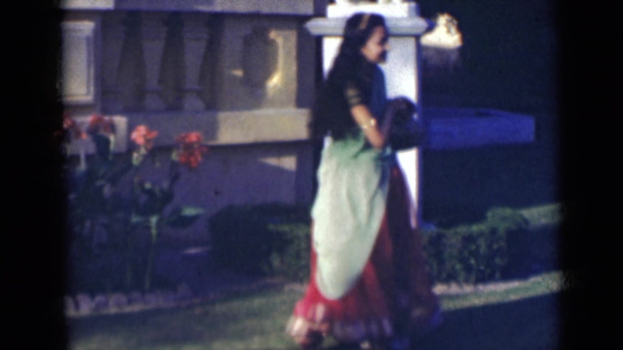 Woman in a green and red sari standing on a lawn near flowers and a pale garden structure.