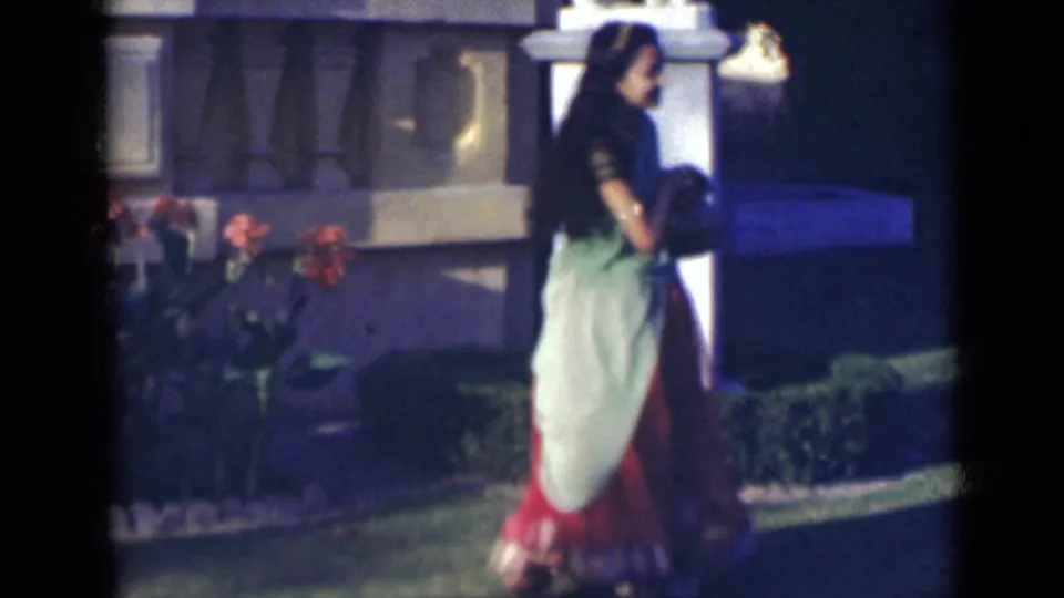 In 1947 India home-movie footage, a woman in a green and red sari stands on grass near red flowers and a pale outdoor structure.