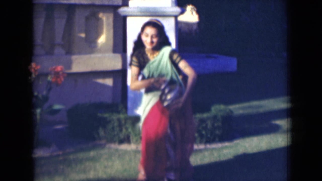 Woman in a green and red sari moving across grass with a jug near a garden structure.