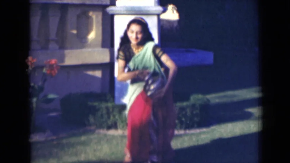 In 1947 India footage, the woman in a green and red sari moves across a lawn holding or carrying a gold-colored jug.