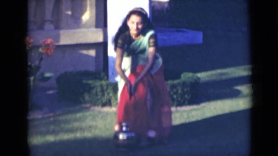 The woman bends on the garden lawn in 1947 India footage, lowering a gold-colored jug toward the grass.