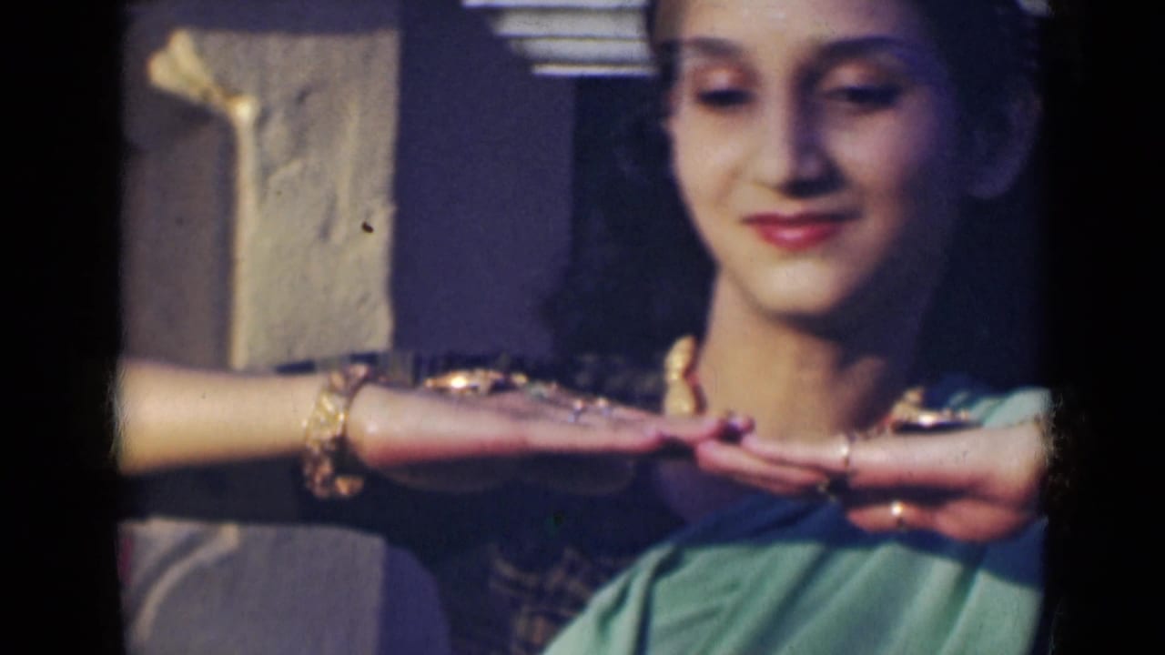 The woman in a green sari holds both hands near her chin in a poised dance gesture, with jewelry visible on her wrists and neck.