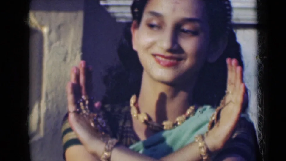 A smiling woman in a green sari and gold-toned jewelry frames her face with raised hands during a traditional dance gesture.
