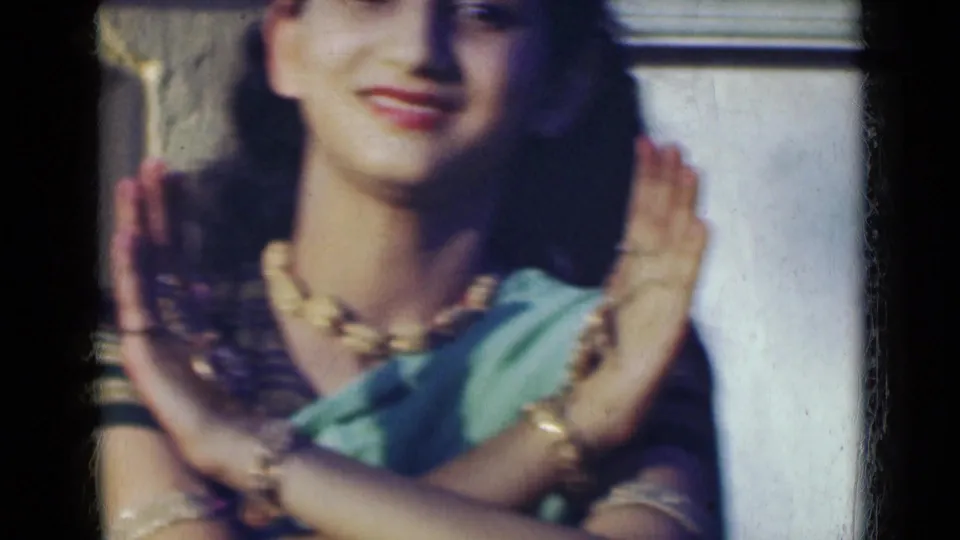 The woman smiles and crosses her wrists in front of her chest, her green sari and gold-toned jewelry visible in soft 8mm color.