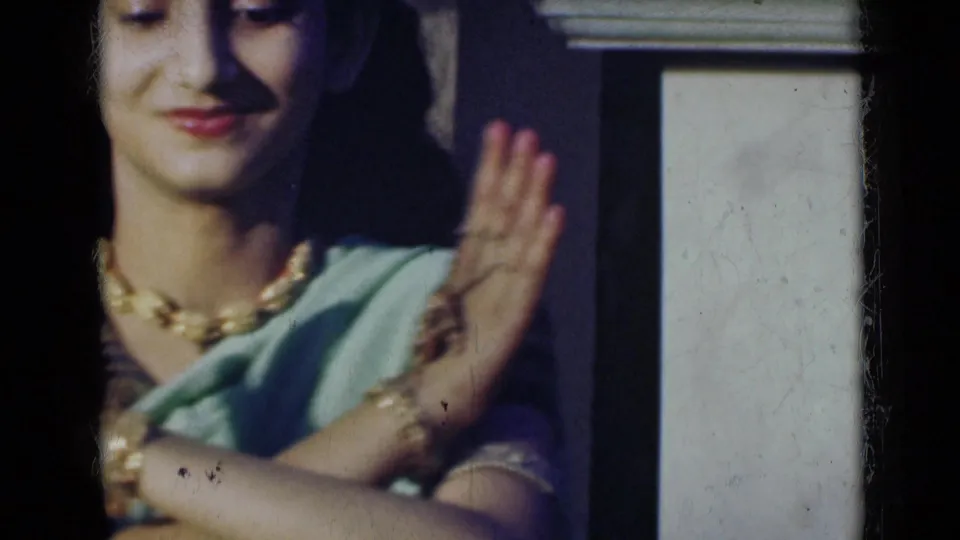 In 1947 India home-movie footage, a young woman in a light green sari and gold-toned jewelry raises one hand in a dance gesture.