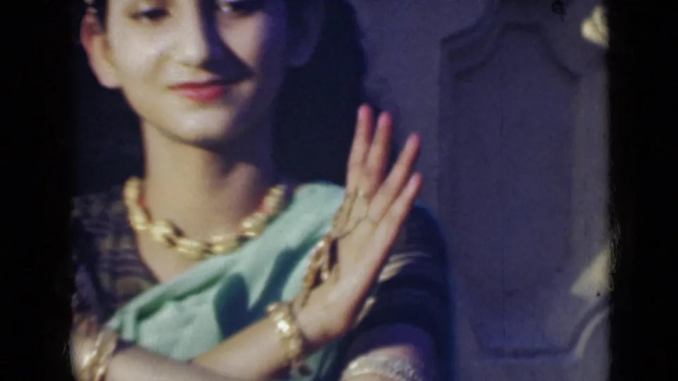 The performer in a light green sari and jewelry holds her hands near her shoulder in a poised dance movement.