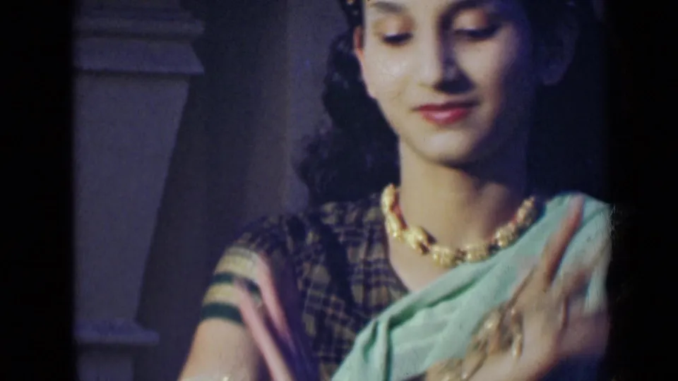 A young woman in sari and jewelry looks downward as one hand moves across her chest in the 8mm dance footage.