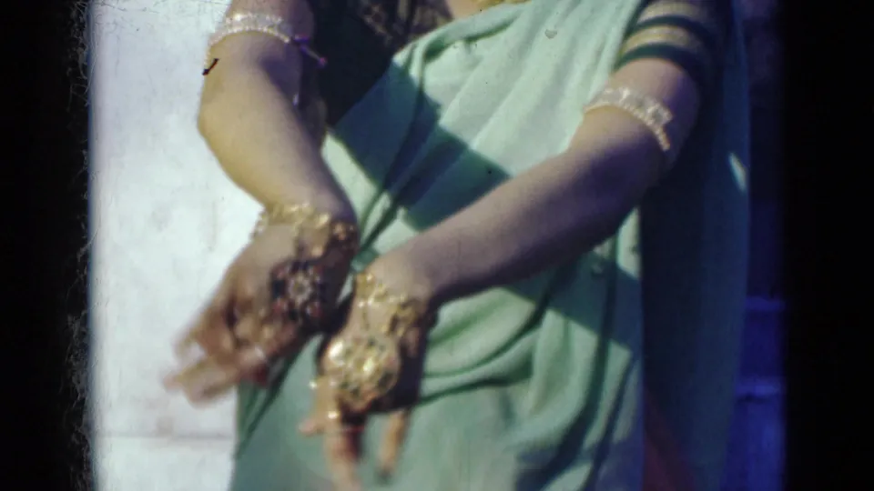 Close framing emphasizes the dancer's bangled and ringed hands gesturing over the folds of a light green sari.