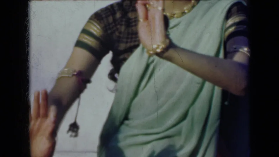 1947 India 8mm frame showing a woman in a light green sari and patterned blouse raising her hands, with bangles and rings visible.