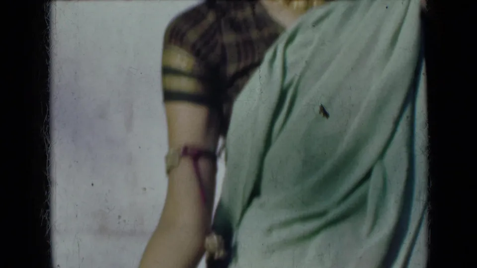 1947 India 8mm frame showing the woman's sari and patterned blouse in a cropped torso view, with one arm lowered and jewelry visible.