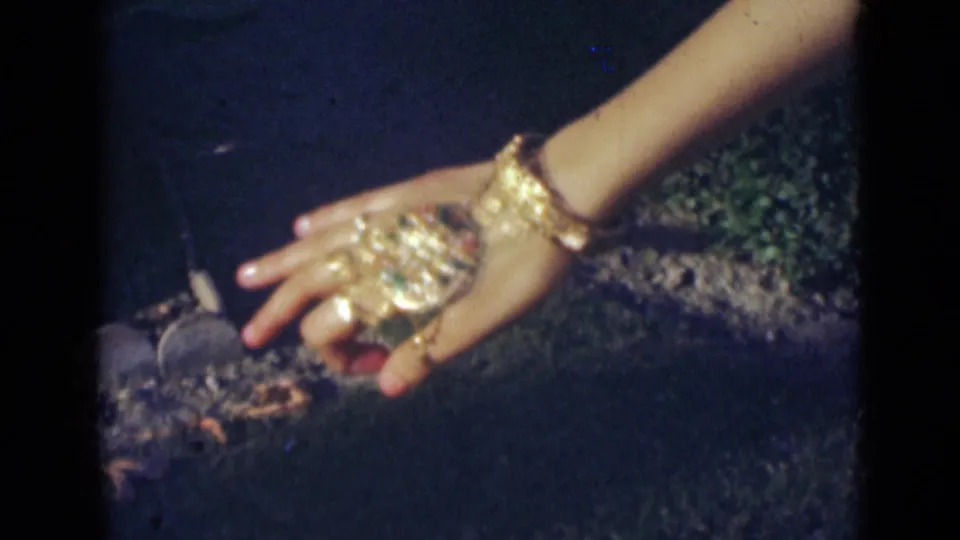 1947 India home-movie frame showing a dancer's hand with rings, bangles, and a large gold ornament lowered near a dark surface.
