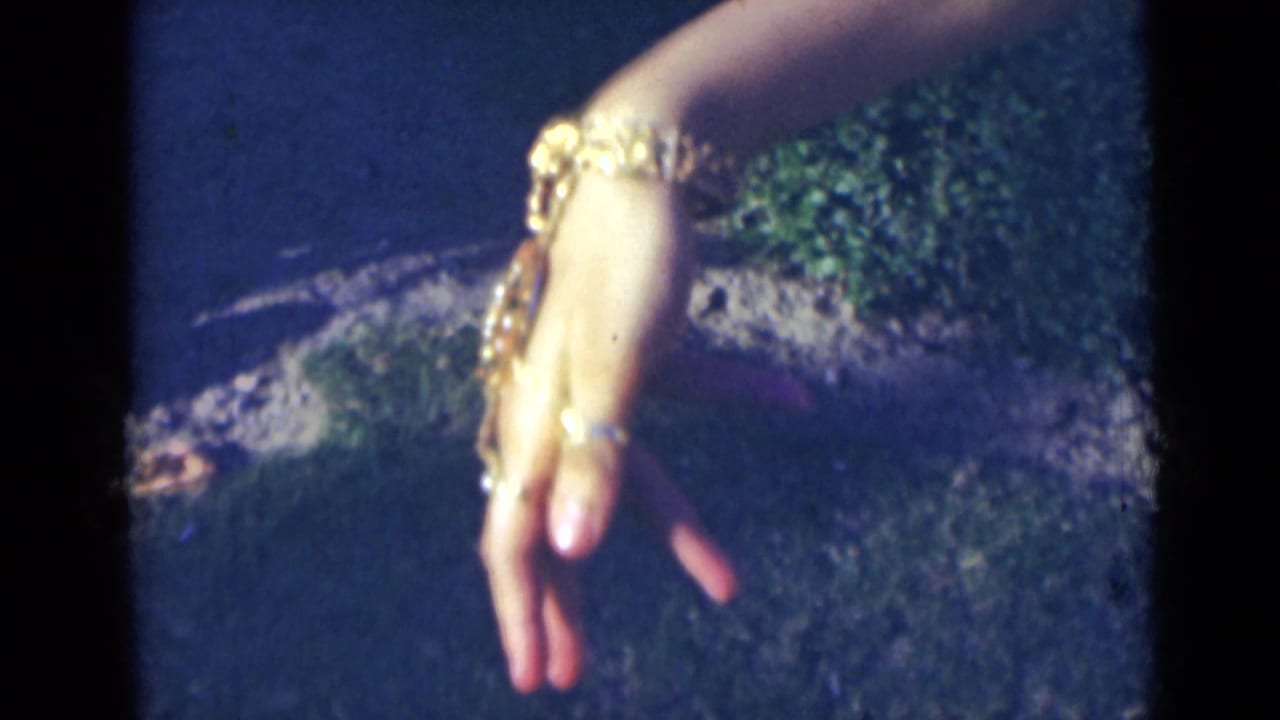 A woman's decorated hand angles downward, showing stacked gold-colored bracelets and rings in the 1947 India home-movie frame.