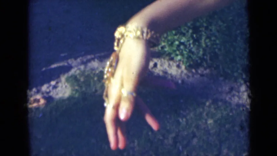 A woman's decorated hand angles downward, showing stacked gold-colored bracelets and rings in the 1947 India home-movie frame.