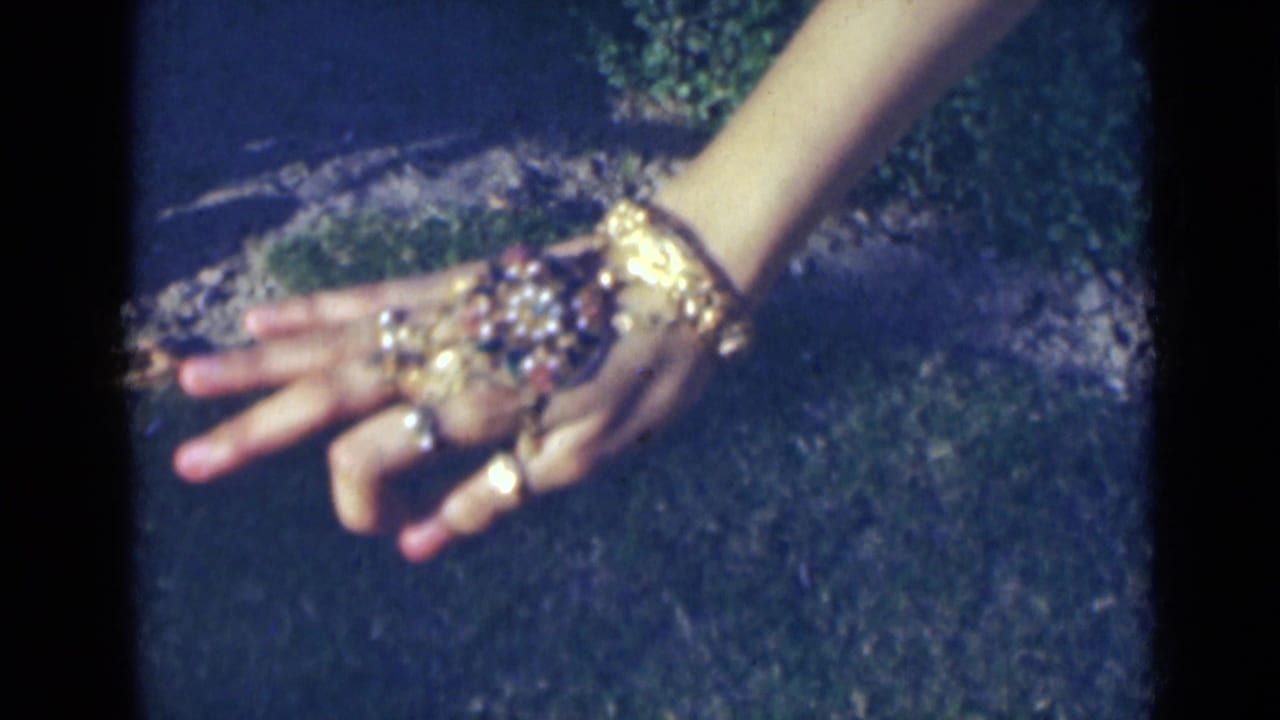 Close view of a woman's hands in India, 1947, showing rings and ornate gold-toned jewelry against dark grass.