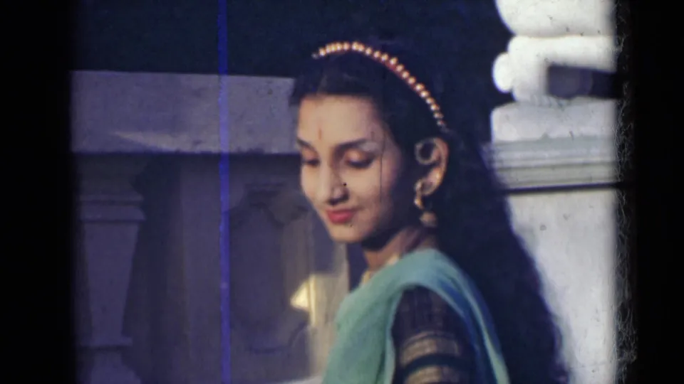 In 1947 India home-movie footage, an adult woman in a green sari and jewelry looks downward near a white stone structure.